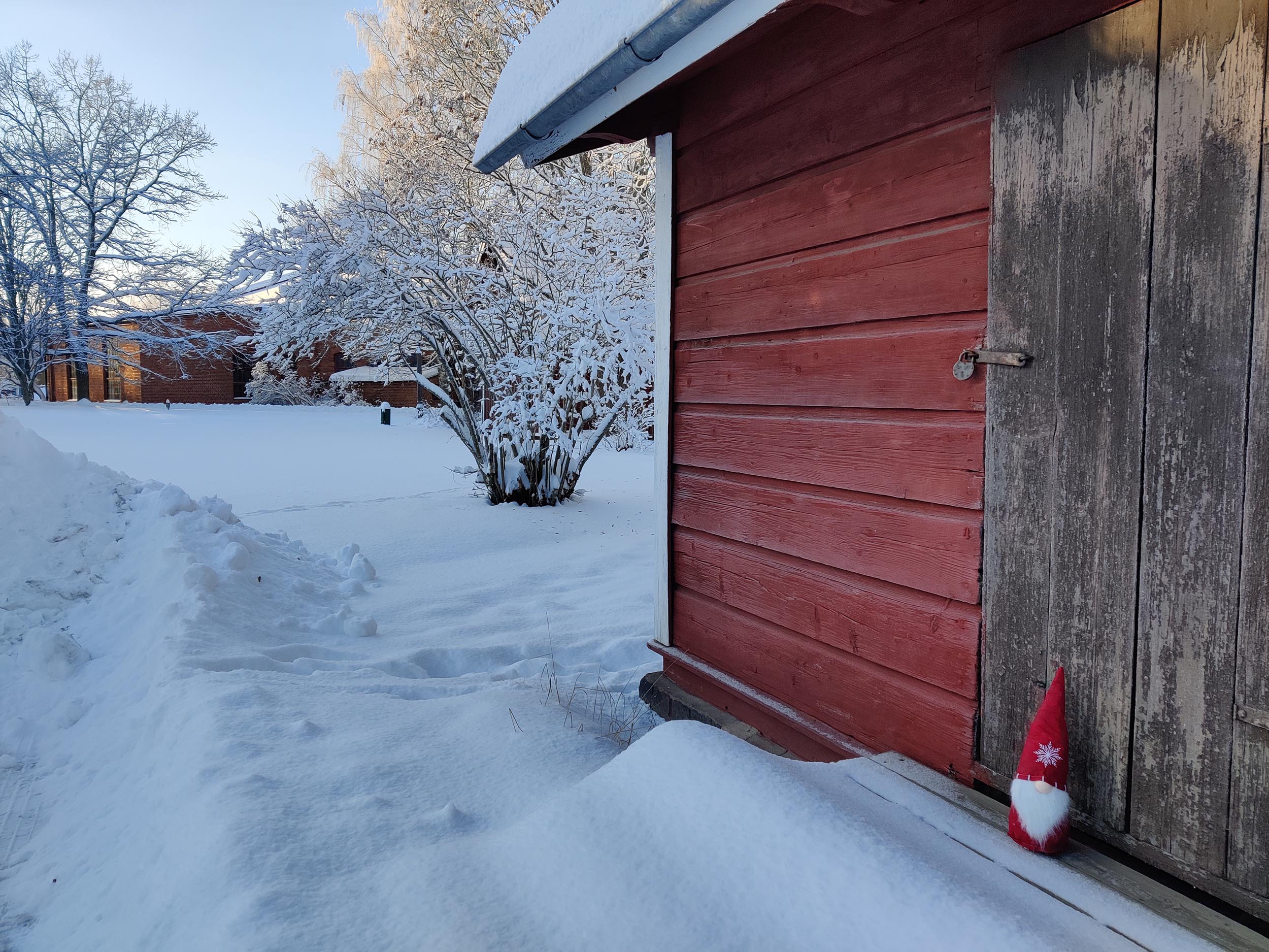 Punainen joulutonttu nököttää rautatierakennuksen edessä museon lumisessa puistossa.