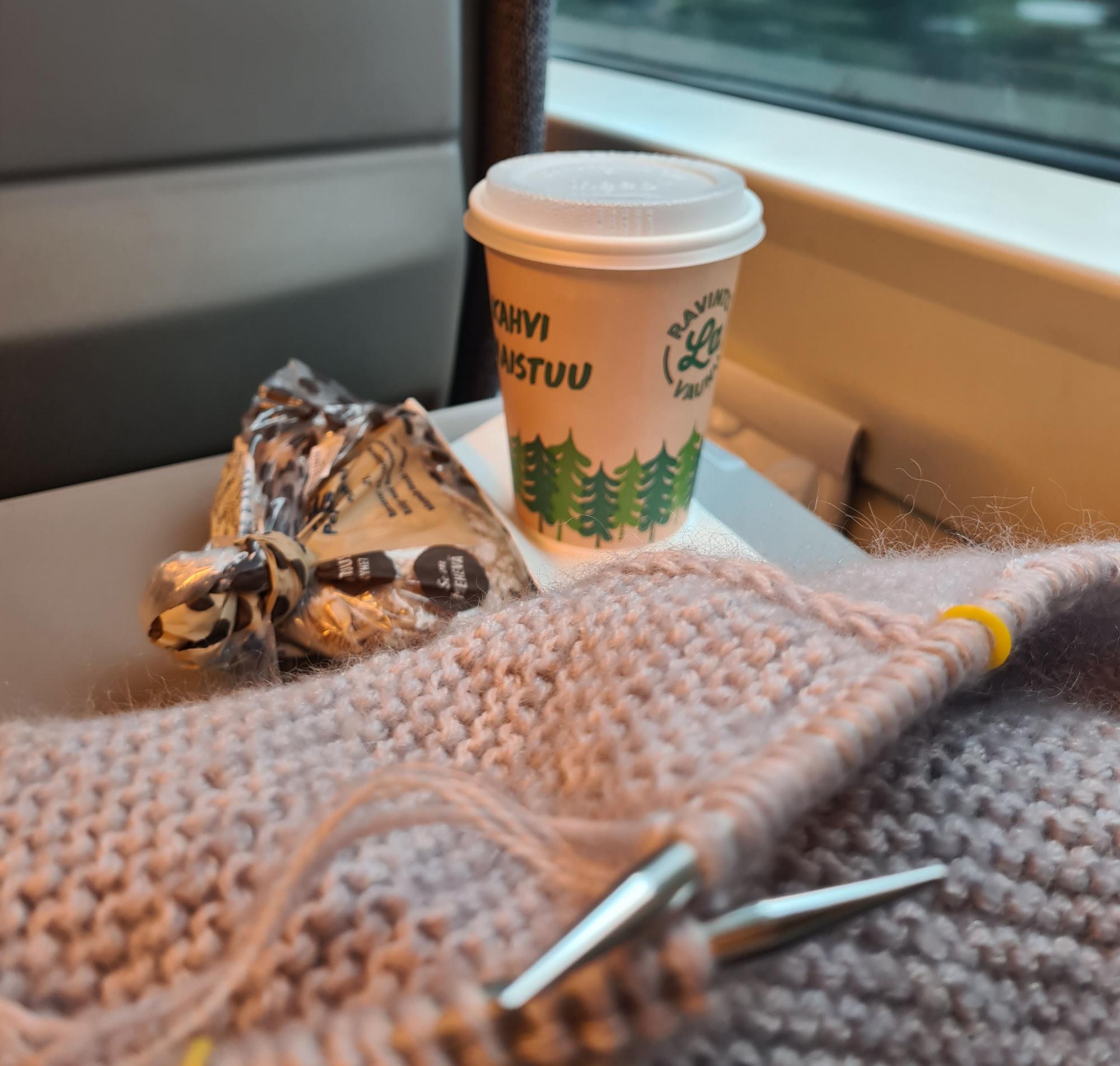 Neuletyö, eväsleipä pussissa ja kahvimuki junan pöydällä. Taustalla junan ikkkuna ohi vilisevä maisema.