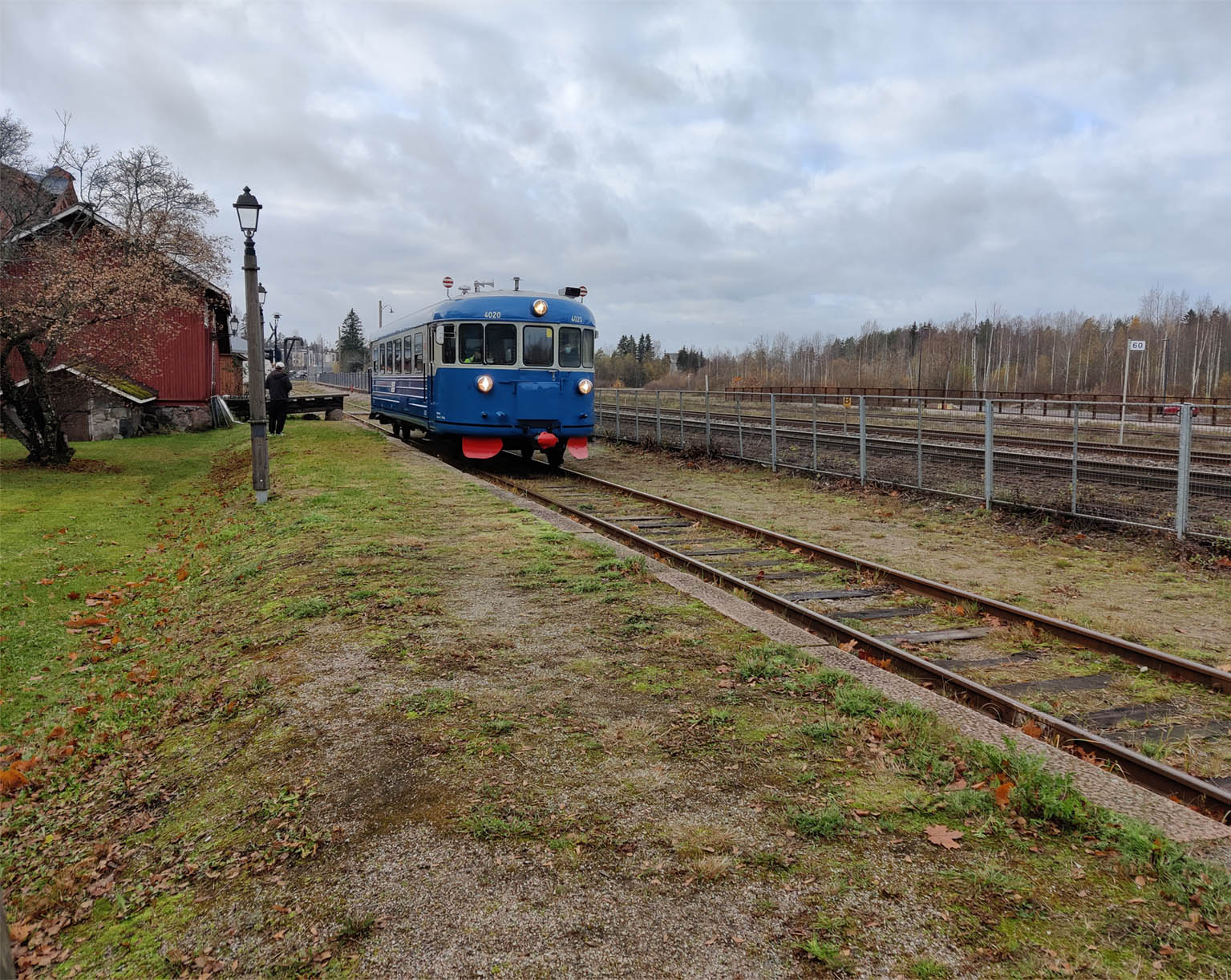 Sininen kiskobussi raiteilla syksyisessä maisemassa punaisen puurakennuksen vieressä.