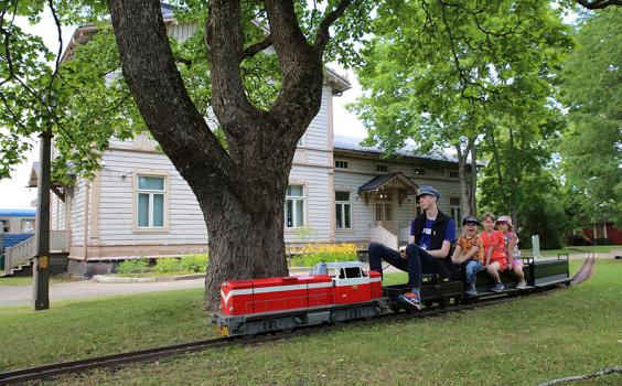 Rautatiemuseon puistojuna liikkeellä museon puistossa kyydissään veturinkuljettaja ja kolme lasta.